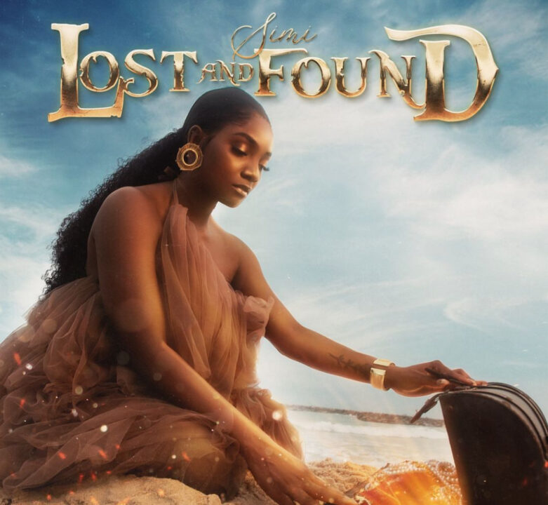 The image is a cover art for a song titled Lost and Found by Simi. It features Simi sitting on a beach, dressed in a flowing, sheer, brown gown. She is looking down and gently touching a treasure chest that is partially buried in the sand, glowing with a golden light. The background is a serene, cloudy sky with the ocean in the distance. The title 